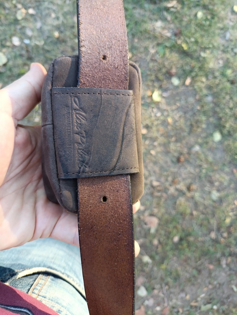 Peut inclure: Une pochette de ceinture en cuir marron fonc&eacute; dot&eacute;e d'une boucle rectangulaire en cuir sur le devant. La boucle est gaufr&eacute;e avec le texte Alex Plus et comprend une sangle en cuir verticale qui la traverse. Le cuir a une finition mate et textur&eacute;e.