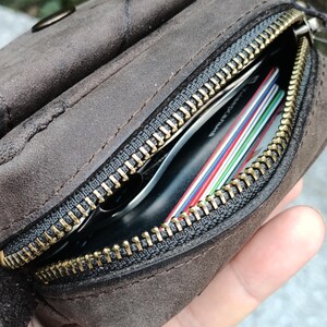 Peut inclure: Une pochette en daim marron fonc&eacute; dot&eacute;e d'une fermeture &eacute;clair en m&eacute;tal couleur laiton. Le compartiment ouvert r&eacute;v&egrave;le plusieurs cartes de cr&eacute;dit empil&eacute;es aux bords color&eacute;s et un objet noir rang&eacute; dans la poche principale de l'accessoire.