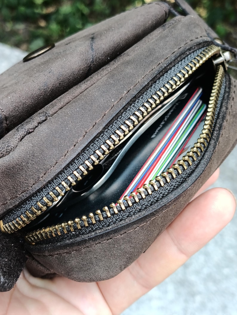 Peut inclure: Une pochette en daim marron fonc&eacute; dot&eacute;e d'une fermeture &eacute;clair en m&eacute;tal couleur laiton. Le compartiment ouvert r&eacute;v&egrave;le plusieurs cartes de cr&eacute;dit empil&eacute;es aux bords color&eacute;s et un objet noir rang&eacute; dans la poche principale de l'accessoire.