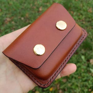 May include: A rectangular brown leather card holder featuring a flap closure secured by two polished gold-tone metal snap buttons. The edges of the leather are stitched with dark thread, creating a defined border around the entire accessory.