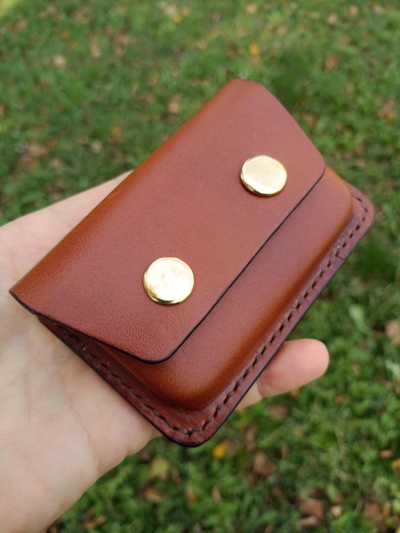 May include: A rectangular brown leather card holder featuring a flap closure secured by two polished gold-tone metal snap buttons. The edges of the leather are stitched with dark thread, creating a defined border around the entire accessory.