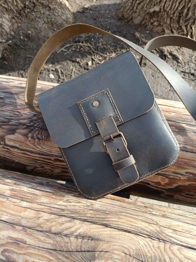 Pu&ograve; includere: Una borsa a tracolla in pelle marrone scuro con chiusura a fibbia e patta. La borsa ha un'unica tracolla ed &egrave; appoggiata su una superficie di legno.