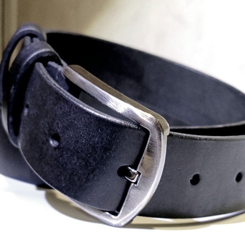 May include: A close-up of a black leather belt with a silver buckle. The belt is curved, showing the texture of the leather and the details of the buckle's design. The buckle is a classic, rectangular shape.
