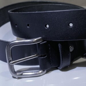 Op de afbeelding: Een zwarte leren riem met een zilverkleurige gesp. De riem heeft verschillende gaten voor verstelling en een klassiek, minimalistisch ontwerp. De rechthoekige gesp heeft een enkele pin.