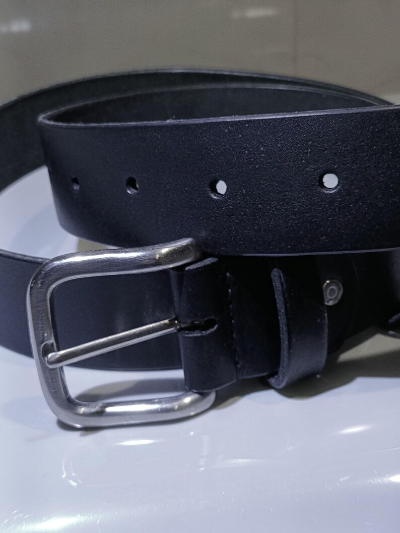 Op de afbeelding: Een zwarte leren riem met een zilverkleurige gesp. De riem heeft verschillende gaten voor verstelling en een klassiek, minimalistisch ontwerp. De rechthoekige gesp heeft een enkele pin.
