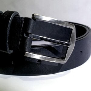 May include: A close-up of a black leather belt with a silver buckle. The belt is rolled up, showing the buckle and the belt loop. The buckle is rectangular with a brushed metal finish.
