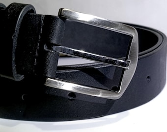 Puede incluir: Primer plano de un cinturón de cuero negro con hebilla plateada. El cinturón está enrollado, mostrando la hebilla y la trabilla. La hebilla es rectangular con acabado metálico cepillado.