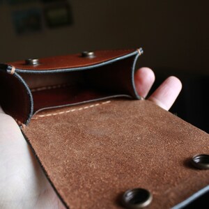 May include: A brown leather pouch with a flap and two metal snaps. The pouch is open and the inside is lined with brown suede.