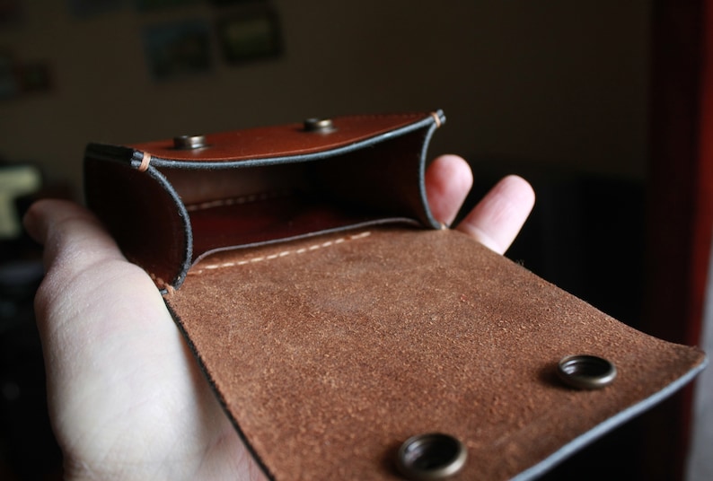 May include: A brown leather pouch with a flap and two metal snaps. The pouch is open and the inside is lined with brown suede.
