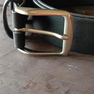 Puede incluir: Un cintur&oacute;n negro con un acabado mate liso y una hebilla de metal plateado pulido. La correa del cintur&oacute;n est&aacute; asegurada por una trabilla de cuero negro a juego, y la hebilla tiene un marco rectangular cl&aacute;sico con un perno redondeado.