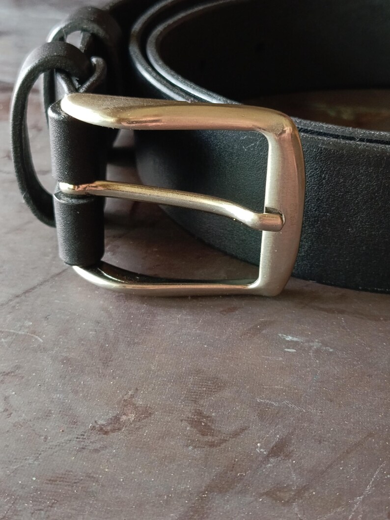 Puede incluir: Un cintur&oacute;n negro con un acabado mate liso y una hebilla de metal plateado pulido. La correa del cintur&oacute;n est&aacute; asegurada por una trabilla de cuero negro a juego, y la hebilla tiene un marco rectangular cl&aacute;sico con un perno redondeado.