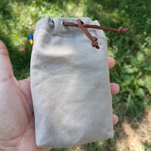May include: A rectangular beige canvas drawstring pouch featuring a brown leather cord closure threaded through silver-toned metal grommets. A small blue and yellow fabric tag is visible on the side seam of the textured, woven fabric bag.