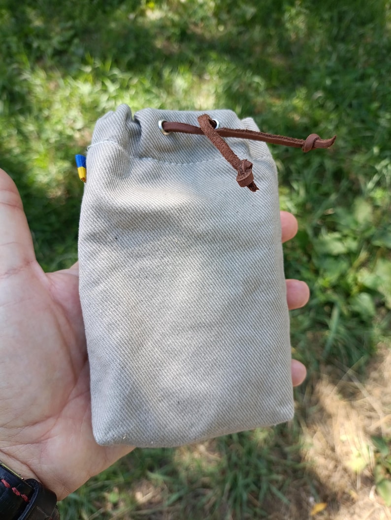 May include: A rectangular beige canvas drawstring pouch featuring a brown leather cord closure threaded through silver-toned metal grommets. A small blue and yellow fabric tag is visible on the side seam of the textured, woven fabric bag.
