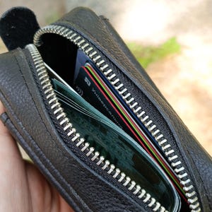 Puede incluir: Un estuche de cuero negro con cremallera y superficie texturizada. El estuche est&aacute; parcialmente abierto, revelando varias tarjetas de cr&eacute;dito y de identificaci&oacute;n guardadas en su interior. La cremallera es plateada con dientes met&aacute;licos.