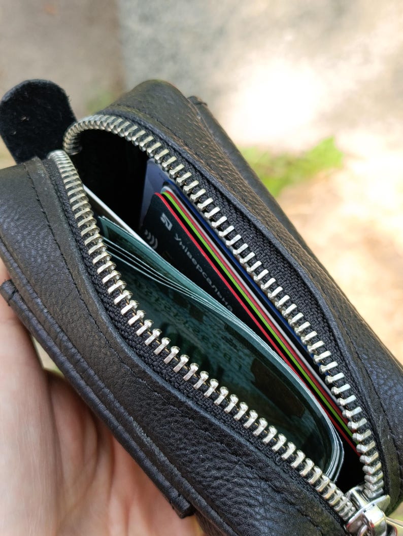 Puede incluir: Un estuche de cuero negro con cremallera y superficie texturizada. El estuche est&aacute; parcialmente abierto, revelando varias tarjetas de cr&eacute;dito y de identificaci&oacute;n guardadas en su interior. La cremallera es plateada con dientes met&aacute;licos.