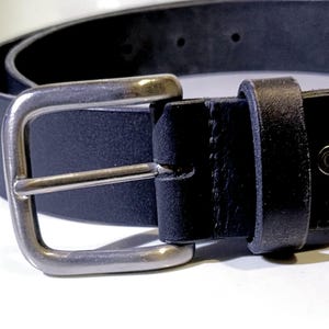 Op de afbeelding: Close-up van een zwarte leren riem met een zilverkleurige gesp. De riem is gemaakt van een glad, donker materiaal en heeft een klassiek rechthoekig gespontwerp. De gesp is gemaakt van glanzend metaal.