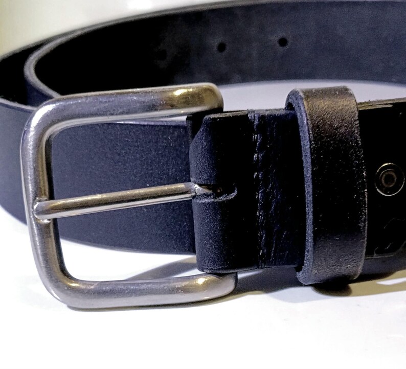 Op de afbeelding: Close-up van een zwarte leren riem met een zilverkleurige gesp. De riem is gemaakt van een glad, donker materiaal en heeft een klassiek rechthoekig gespontwerp. De gesp is gemaakt van glanzend metaal.
