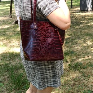 May include: A deep red tote bag with a textured crocodile skin pattern, featuring two vertical shoulder straps attached with gold-toned metal rivets. The bag has a structured rectangular shape and a glossy finish, showcasing a dark red hue.