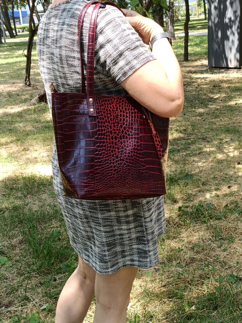 May include: A deep red tote bag with a textured crocodile skin pattern, featuring two vertical shoulder straps attached with gold-toned metal rivets. The bag has a structured rectangular shape and a glossy finish, showcasing a dark red hue.