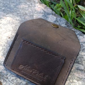 May include: A dark brown leather card holder with a flap closure and a single brass-colored snap button. The front features a stitched pocket embossed with the text Alex Polak HANDMADE LEATHER GOODS in a script and block font combination.
