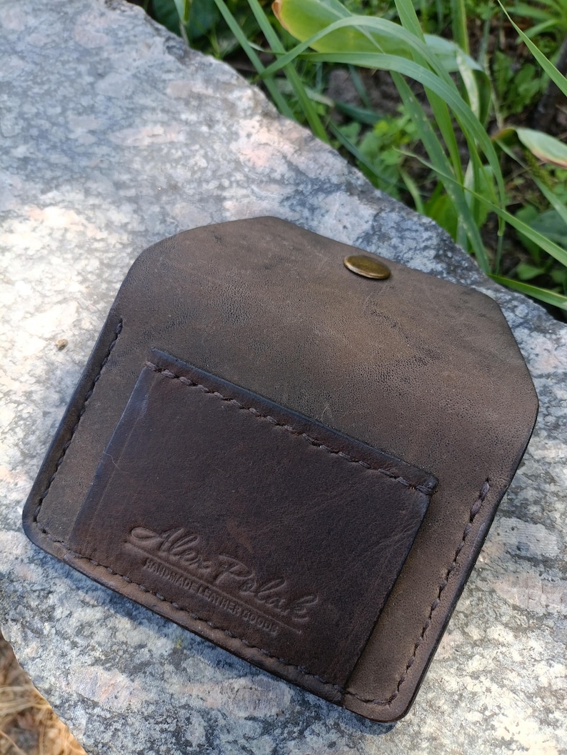 May include: A dark brown leather card holder with a flap closure and a single brass-colored snap button. The front features a stitched pocket embossed with the text Alex Polak HANDMADE LEATHER GOODS in a script and block font combination.