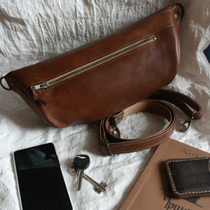 May include: A brown leather fanny pack with a zipper closure and an adjustable strap. The bag is lying on a white fabric surface with a phone, keys, and a wallet.