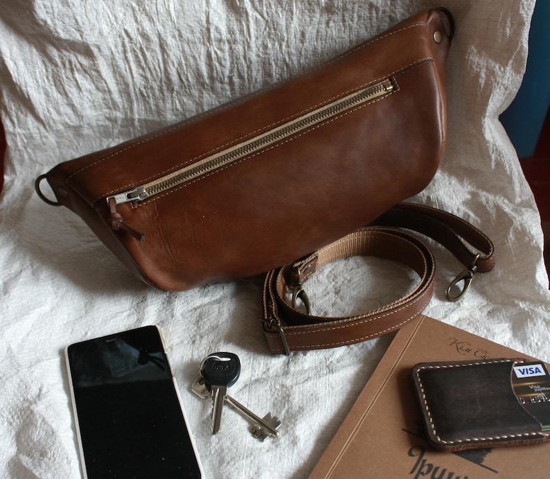 May include: A brown leather fanny pack with a zipper closure and an adjustable strap. The bag is lying on a white fabric surface with a phone, keys, and a wallet.