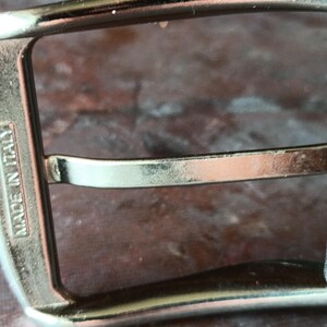 Puede incluir: Una hebilla de cintur&oacute;n de metal plateado con un marco rectangular y un perno central. El lateral del marco de la hebilla tiene grabada la inscripci&oacute;n MADE IN ITALY. La hebilla est&aacute; unida a una correa de cuero negro visible a la derecha.