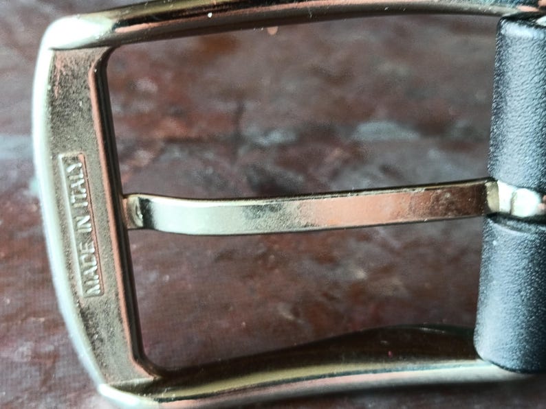 Puede incluir: Una hebilla de cintur&oacute;n de metal plateado con un marco rectangular y un perno central. El lateral del marco de la hebilla tiene grabada la inscripci&oacute;n MADE IN ITALY. La hebilla est&aacute; unida a una correa de cuero negro visible a la derecha.