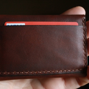 May include: A brown leather card holder with a visible red card inside. The card holder is rectangular, with stitched edges, and appears to be handmade. The leather has a smooth texture and a rich, dark brown color.