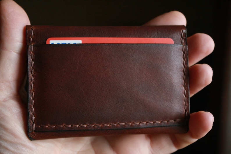 May include: A brown leather card holder with a visible red card inside. The card holder is rectangular, with stitched edges, and appears to be handmade. The leather has a smooth texture and a rich, dark brown color.