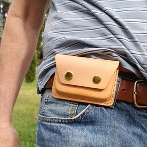 May include: A light tan leather belt pouch featuring a rectangular flap secured by two polished gold-tone metal snap buttons. The pouch is constructed with visible stitching along the edges and is attached to a brown leather belt worn over blue jeans.