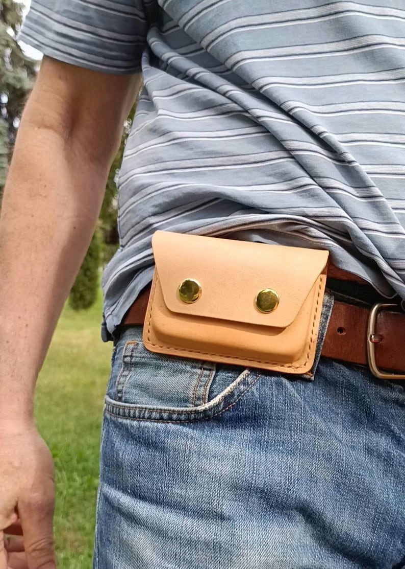 May include: A light tan leather belt pouch featuring a rectangular flap secured by two polished gold-tone metal snap buttons. The pouch is constructed with visible stitching along the edges and is attached to a brown leather belt worn over blue jeans.