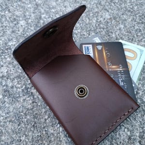 May include: A dark brown leather card holder with a flap closure secured by a brass-toned metal snap button. The interior contains a black credit card and a portion of a one hundred dollar bill. The edges feature visible stitching detail.
