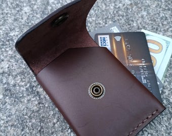 May include: A dark brown leather card holder with a flap closure secured by a brass-toned metal snap button. The interior contains a black credit card and a portion of a one hundred dollar bill. The edges feature visible stitching detail.