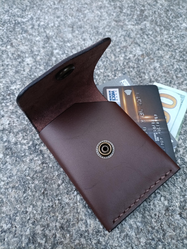 May include: A dark brown leather card holder with a flap closure secured by a brass-toned metal snap button. The interior contains a black credit card and a portion of a one hundred dollar bill. The edges feature visible stitching detail.
