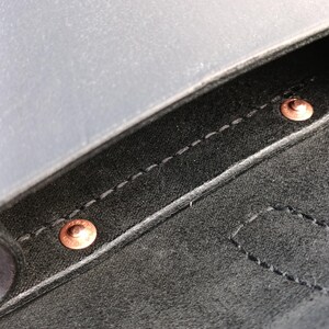 May include: Close-up of a black leather item with copper rivets and white stitching. The leather has a matte finish. The rivets are circular and appear to be holding the leather together. The stitching is visible along the edges.