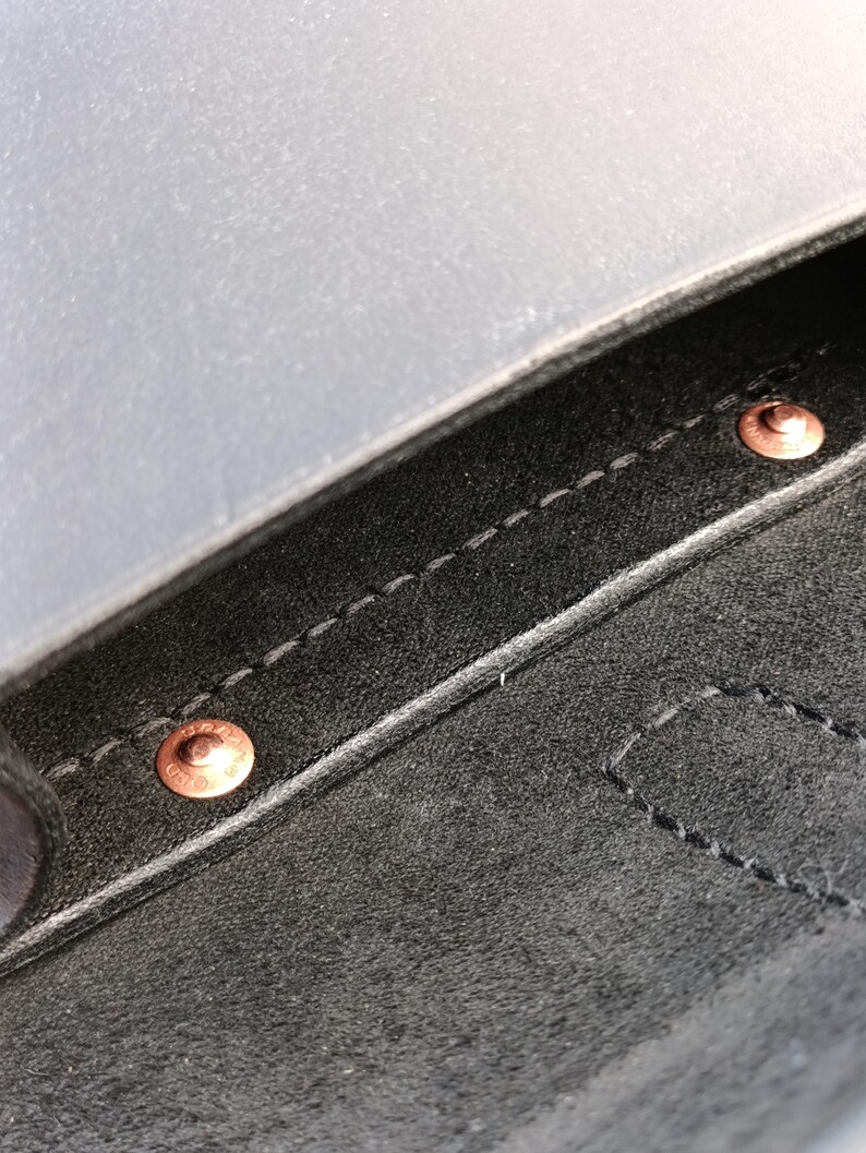 May include: Close-up of a black leather item with copper rivets and white stitching. The leather has a matte finish. The rivets are circular and appear to be holding the leather together. The stitching is visible along the edges.