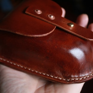 May include: A handcrafted, brown leather case with a curved design. The case features a flap closure secured by a brass stud and two copper rivets. White stitching accents the edges, and the leather has a glossy finish.