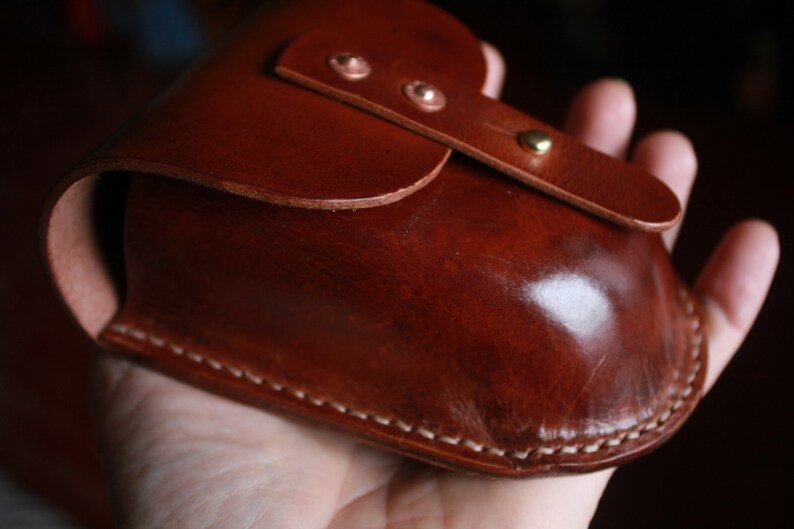May include: A handcrafted, brown leather case with a curved design. The case features a flap closure secured by a brass stud and two copper rivets. White stitching accents the edges, and the leather has a glossy finish.