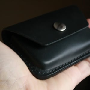 Puede incluir: Una cartera de cuero negro con un borde cosido y un cierre de presi&oacute;n plateado.
