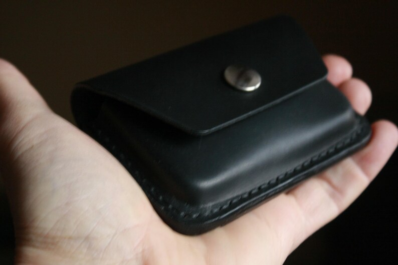 Puede incluir: Una cartera de cuero negro con un borde cosido y un cierre de presi&oacute;n plateado.
