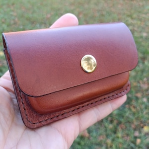 May include: A small, rectangular brown leather card holder or wallet featuring a smooth finish, rounded corners, and a single polished gold-tone metal snap button closure centred on the front flap with visible dark stitching along the edges.