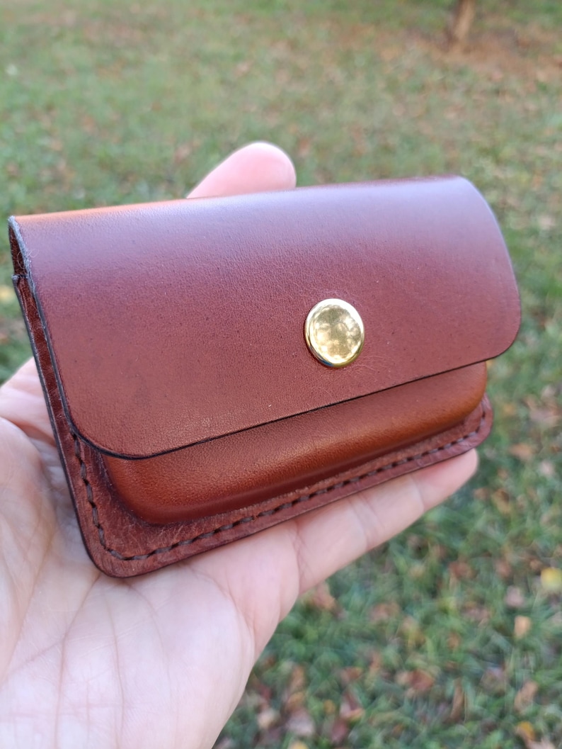 May include: A small, rectangular brown leather card holder or wallet featuring a smooth finish, rounded corners, and a single polished gold-tone metal snap button closure centred on the front flap with visible dark stitching along the edges.