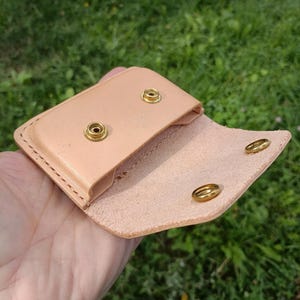 May include: A light tan leather pouch featuring a fold-over flap with two brass snap fasteners. The interior of the pouch also displays two matching brass snap components. The edges are secured with visible stitching along the perimeter of the item.