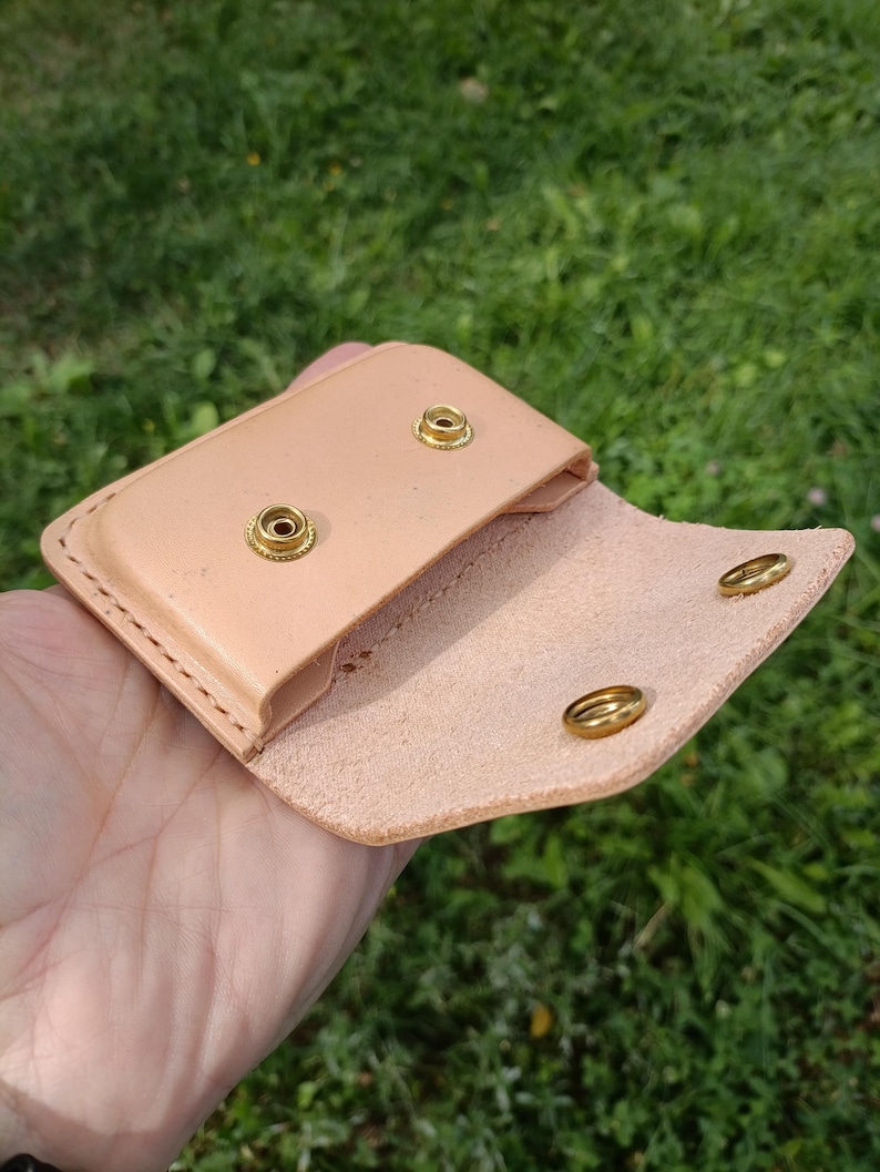 May include: A light tan leather pouch featuring a fold-over flap with two brass snap fasteners. The interior of the pouch also displays two matching brass snap components. The edges are secured with visible stitching along the perimeter of the item.
