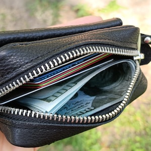 Puede incluir: Una cartera de cuero negro texturizado con cierre de cremallera de metal plateado. La cartera abierta contiene varias tarjetas de cr&eacute;dito de colores y un fajo de billetes de cien d&oacute;lares estadounidenses en el compartimento principal.