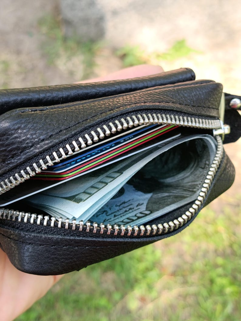 Puede incluir: Una cartera de cuero negro texturizado con cierre de cremallera de metal plateado. La cartera abierta contiene varias tarjetas de cr&eacute;dito de colores y un fajo de billetes de cien d&oacute;lares estadounidenses en el compartimento principal.