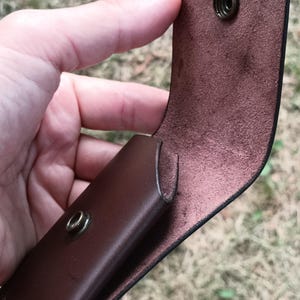 May include: A dark brown leather pouch featuring a smooth exterior and a textured interior. The pouch has a flap closure secured by a circular brass-colored metal snap button, with the corresponding snap component visible on the front body.