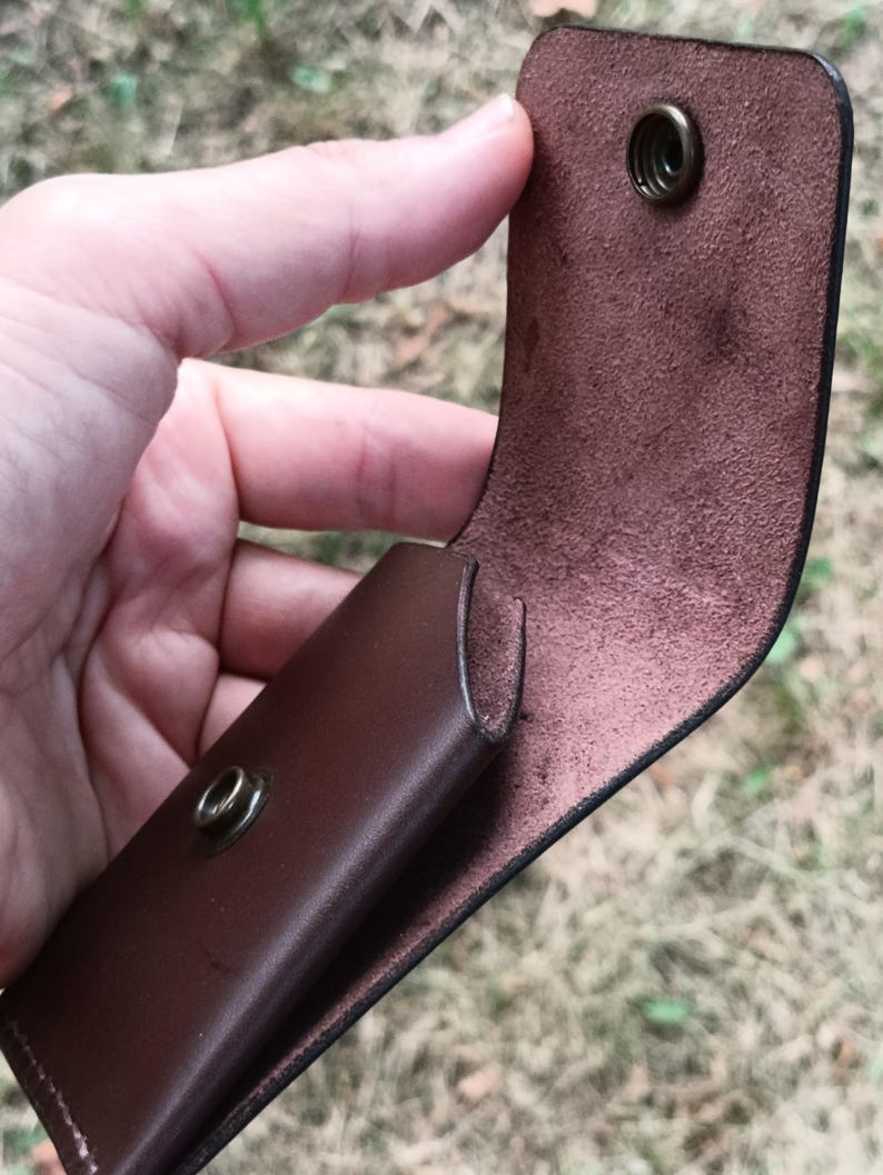 May include: A dark brown leather pouch featuring a smooth exterior and a textured interior. The pouch has a flap closure secured by a circular brass-colored metal snap button, with the corresponding snap component visible on the front body.
